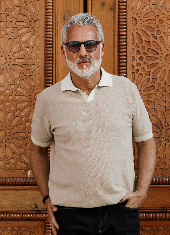 A man with gray hair and a beard, wearing sunglasses and the Buhl Split Neck Knit Polo in beige, stands with one hand in his pocket before an ornate wooden door.