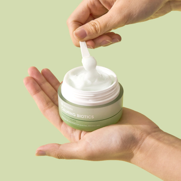 Hands holding a jar of Calming Biotics Blemish Cream | Leaders with a spatula scooping out the smooth white skincare cream against a pale green background