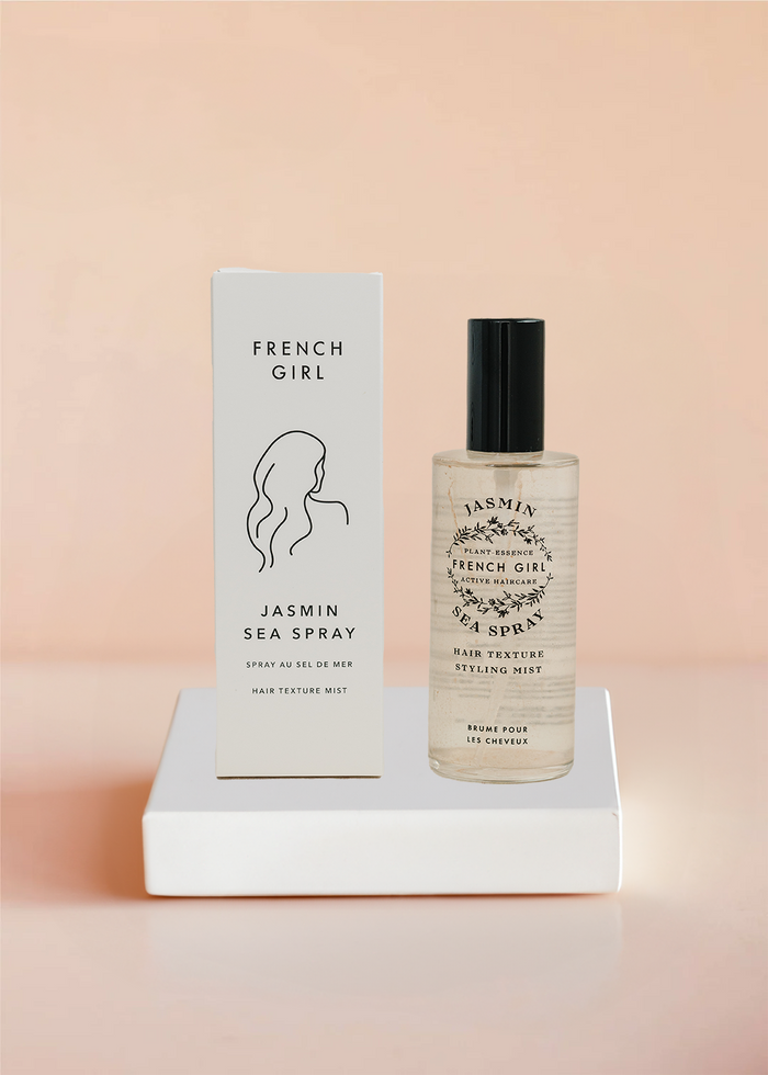 Clear bottle of Jasmin Sea Spray - Hair Texture Mist | French Girl with black cap next to white packaging box featuring minimalist line drawing of a woman’s hair on soft peach background.