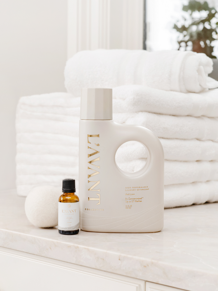 Minimalist white bottle of Laundry Oil | L'AVANT Collective with small amber glass bottle next to it, set on marble countertop with neatly folded white towels in the background.