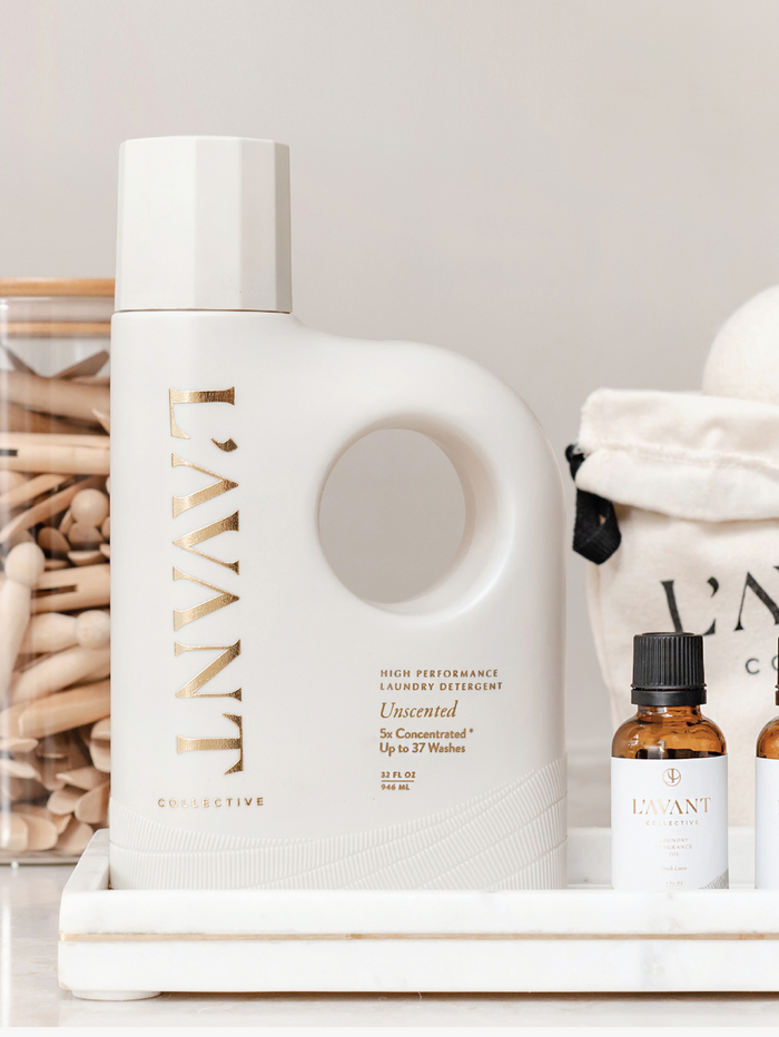Minimalist white bottle of Laundry Oil | L'AVANT Collective with gold lettering, displayed on a white tray alongside small amber glass bottles and soft fabric items.