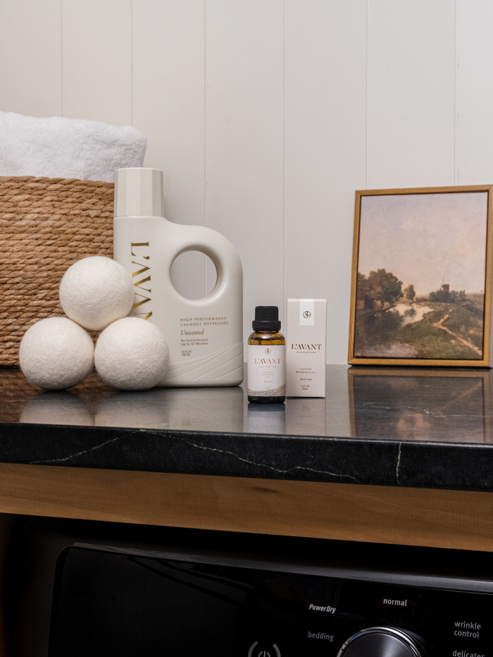 Laundry Oil | L'AVANT Collective displayed on a countertop with natural wool dryer balls, a basket with towels, and a framed landscape painting in a cozy laundry room setting.