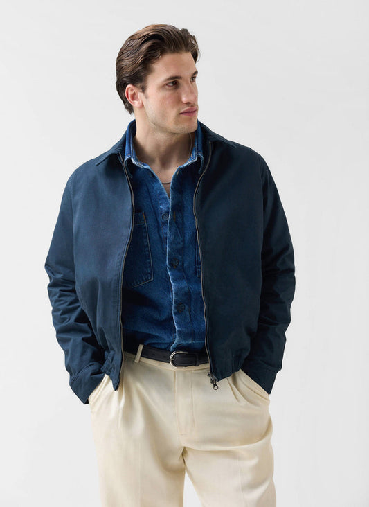A man models the Percival Leo Harrington jacket in navy, styled with a denim shirt and cream trousers.