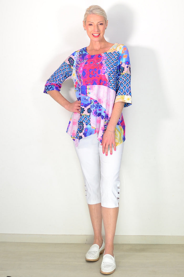 A woman modeling a vibrant, multicolored patterned tunic top with three-quarter sleeves, paired with white capri pants and white slip-on shoes, standing against a plain white wall.
