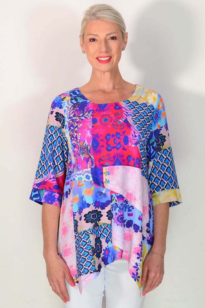 A woman modeling a vibrant, multicolored patchwork-patterned blouse with three-quarter sleeves and a scoop neckline, paired with white pants against a plain white background.