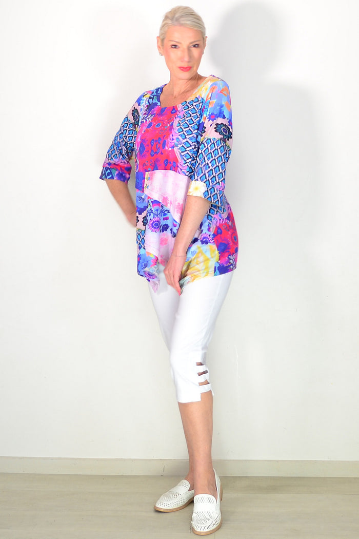 A woman modeling a vibrant, multicolored short-sleeve top with a mix of geometric and floral patterns in blue, pink, purple, and yellow, paired with white capri pants and white woven slip-on shoes, standing against a plain white wall.