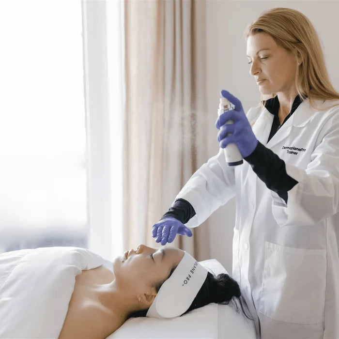 A skincare professional wearing gloves sprays Satisfaction Mist | DermaplanePro on a relaxed woman's face in a bright, calming treatment room.