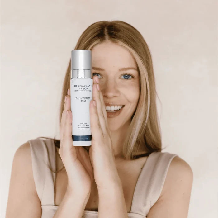Young woman with clear skin smiling and holding a bottle of Satisfaction Mist | DermaplanePro skincare product in front of her face against a neutral background