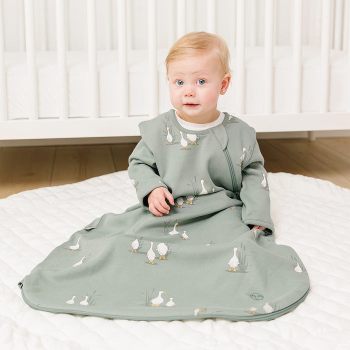 Baby in a green sleeping bag with sleeves with a duck pattern sitting on a baby rug.
