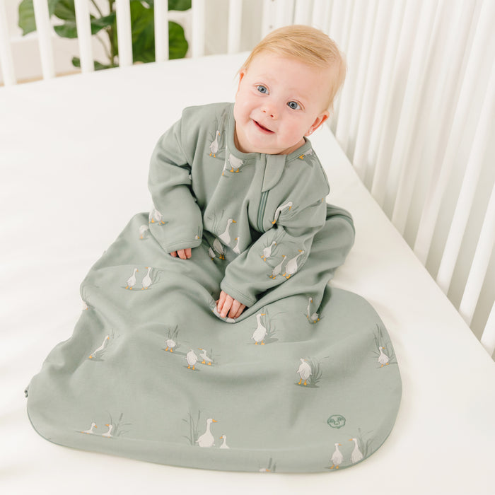 Baby in a green sleeping bag with sleeves featuring a goose pattern, sitting in a crib.